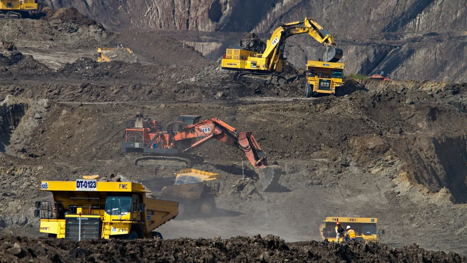 An active open-pit mine: excavators and haul trucks working the benches, with two workers in high-visibility gear on the floor